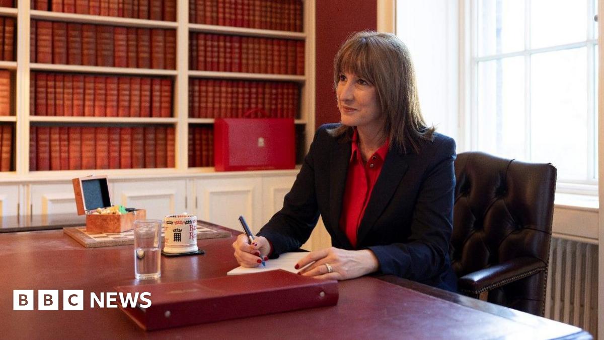 Rachel Reeves sits at a desk in the Treasury, holding a pen. On the desk in front  of her is a red folder, water, a mug and a chess set. She has a red box behind her. She is dressed in a red shirt and black jacket.
