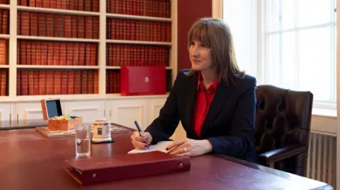 Kirsty O'Connor / Treasury Rachel Reeves sits at a desk in the Treasury, holding a pen. On the desk in front  of her is a red folder, water, a mug and a chess set. She has a red box behind her. She is dressed in a red shirt and black jacket.