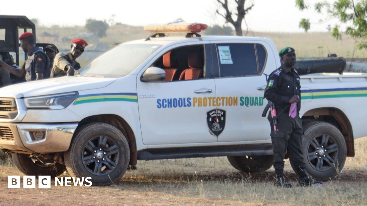 A police officer stands next to a white pickup truck that has the words 'schools protection squad' on the side of it, with other security officials behind the truck which is parked on a grassy area next to a tree in Kebbi, Nigeria on 17 November.