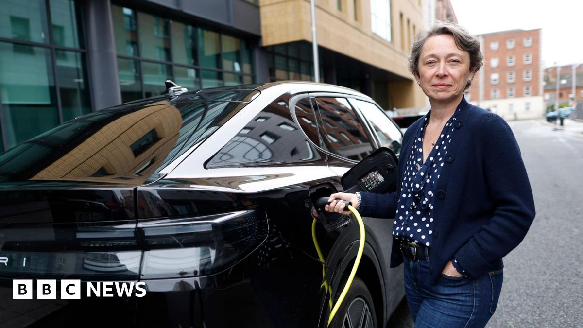 Ford UK's managing director Lisa Brankin wearing blue jeans, a navy blue blouse with white polka dots and a navy blue cardigan. She is standing next to a black Ford Capri connecting it to an electric charger and smiling at the camera