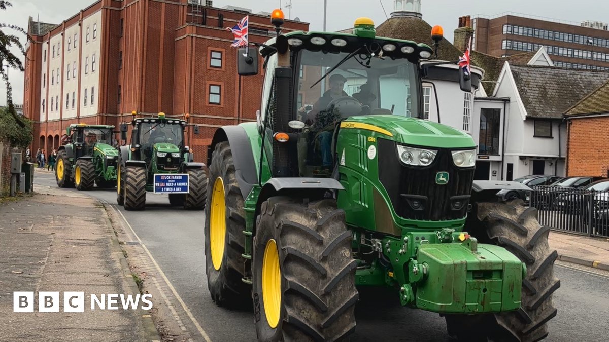 A green tractor with British flags on either side of the driver's cab moves through a town centre road. Several other tractors of similar colour and style follow it from behind. Buildings surround the road and there is a car park to the right hand side.