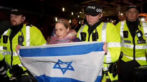 PA Media A women named Emily carrying an Israel flag is moved away by police officers from pro Palestine campaigners, who are protesting on Trinty Road outside Villa Park.