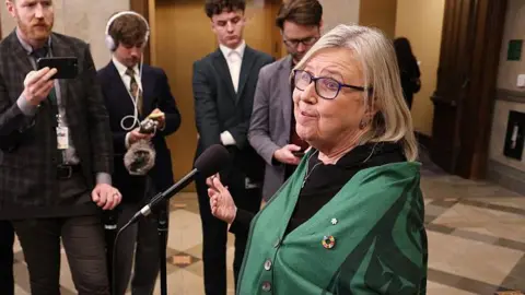 Getty Images Canada's Green Party leader Elizabeth May speaks to the media after attending Question Period on Parliament Hill on November 17, 2025 in Ottawa, Canada. She is speaking outside the House of Commons at a microphone and is surrounded by reporters who have their phones and recorders out. She has short, shoulder-length straight grey hair and is wearing a black top with a forest green buttoned poncho. 