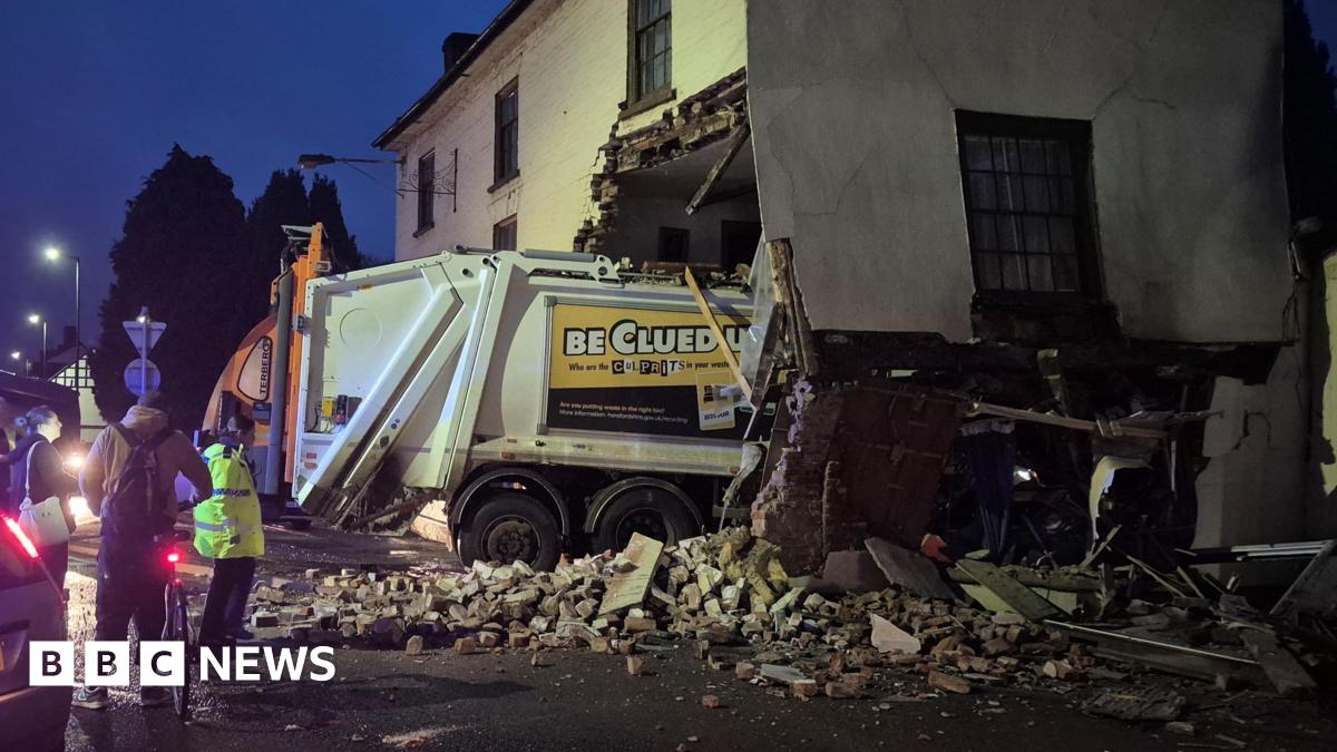 A bin lorry has crashed through the ground floor of a building, with debris lying around the area beside it. There is a silver Ford car in the foreground and a number of people gathered nearby the scene.