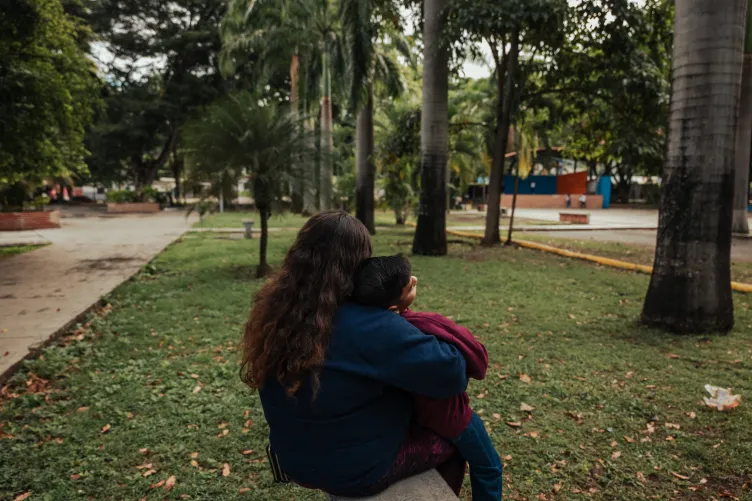 A woman and child wearing blue and maroon sweatsuits sit on a beach facing away from the camera in a grassy area of a city park, with tall palm trees and other lush tropical plants.