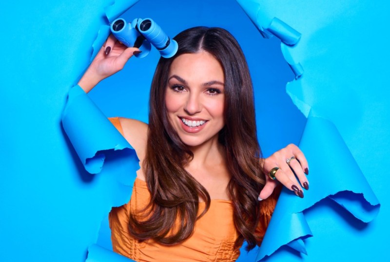 Woman with long hair smiling and holding blue binoculars, emerging from a torn blue paper background, wearing an orange top and displaying stylish rings.