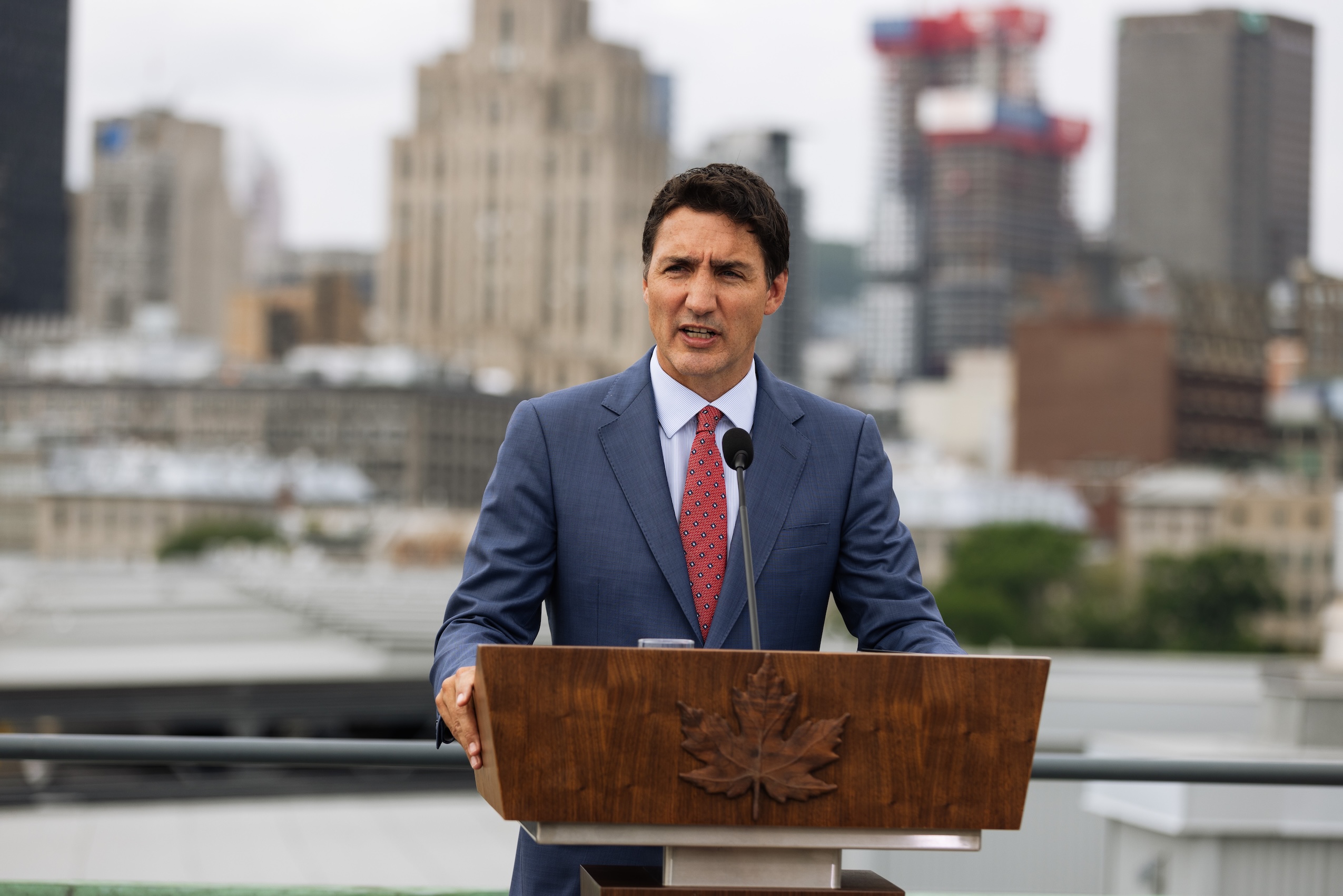 Justin Trudeau speaking at a podium with city buildings in the background.
