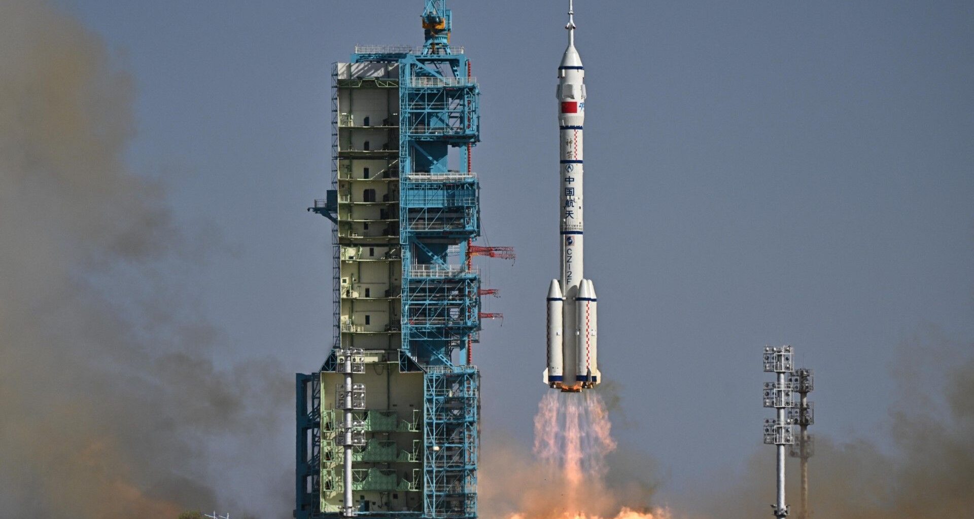 a white rocket with four small boosters at its base lifts off above a plume of fire beside a blue launch tower