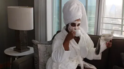 Tawana Musvaburi A woman is sat on a sofa in a white bath robe, having a cup of tea whilst wearing a face mask and reading the newspaper. 