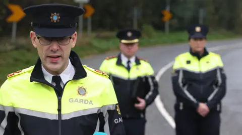 PA Media Supt Charlie Armstrong speaking to the media near the scene, there are two officers standing behind him. He is wearing a garda uniform with navy hat and galsses.