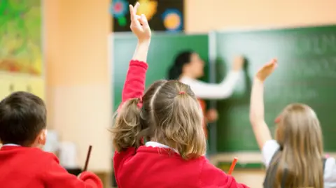 Getty Images Primary school children are sitting at desks in their classroom. Their uniform is a red jumper. One girl is wearing a grey dress. A teacher is writing with chalk on the board. A girl with pigtails has her hand up.