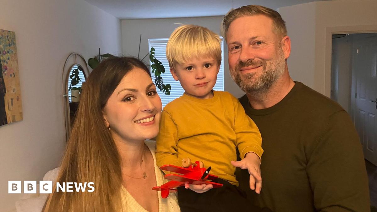 Monika, Henry and Leo smile at the camera in a living room. Monika, who has long brown hair, and Henry who has blonde short hair and a brown jumper are holding toddler Leo who is holding a red toy plane and has a yellow jumper on.
