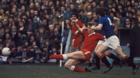SNS Willie Young wearing a red Aberdeen shirt tackling a Rangers player wearing a blue shirt with white shorts and black socks.