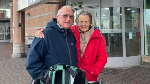Elderly married couple David and Anne Patterson stand next to each other with their arms around their shoulders while they stand outside Jersey Airport main terminal. David has glasses on and a navy blue coat while Anne has a red coat on with a beige undercoat. A green and black rucksack is on top of a luggage trolley in front of them.