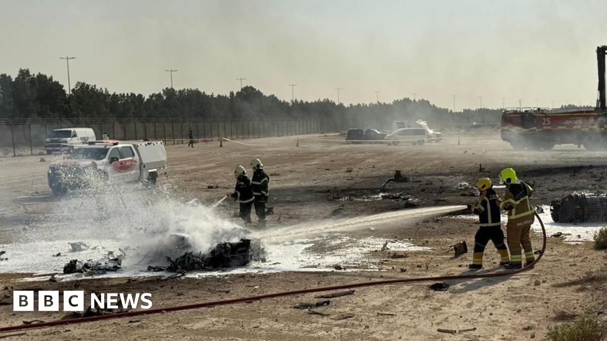 Firefighters work at the scene of a plane crash, water spraying onto a burnt piece of metal on the ground, it is in an open stretch of land with cars and a fence visible in the background.