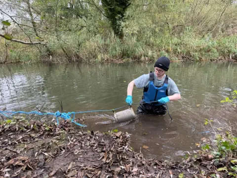 Daniel Jolly/University of East Anglia Daniel Jolly stands in a river holding small cages which contain wet wipes for his experiment. 