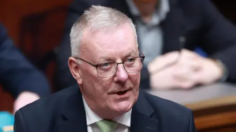 PA Media Nesbitt has short grey hair, black metal glasses and is speaking in the picture. He is wearing a black blazer, green tie and white shirt.