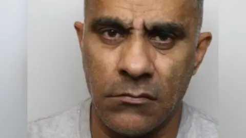 South Yorkshire Police A man with short grey hair and brown eyes. He is wearing a grey t-shirt and is in front of a grey background. It is a custody photo.