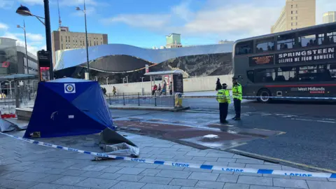 BBC  blue police tent on a pavement, cordoned off by blue and white police tape. Two police officers are standing inside the tape wearing black hats and yellow hi-vis jackets. a bus drives past