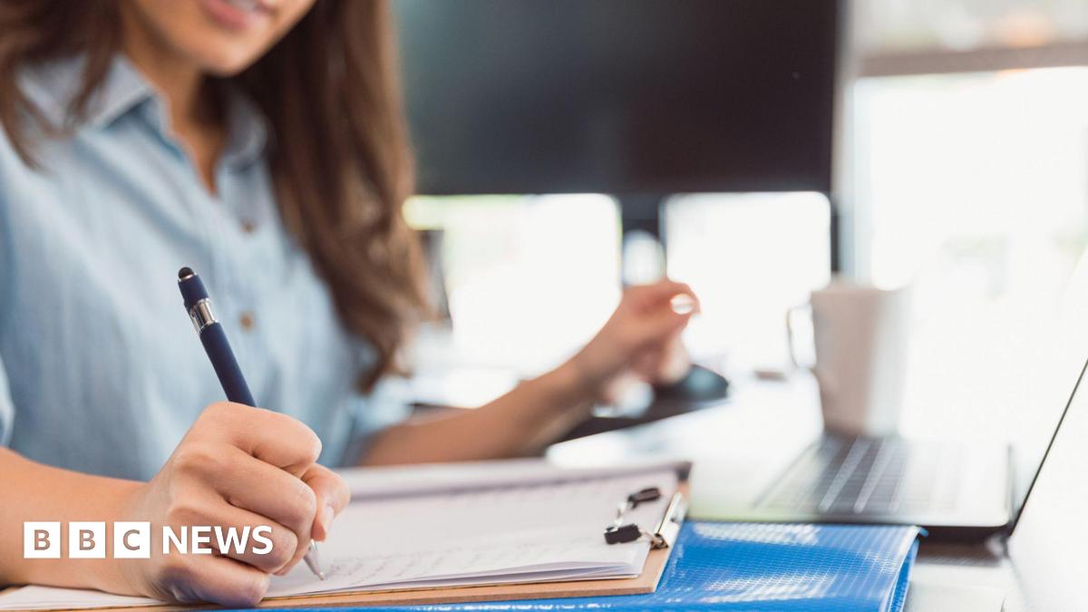 A woman sitting at a desk, writing on a clipboard with a pen. A blue folder is underneath the clipboard and a laptop and white coffee mug are visible in the background on the desk. She is wearing a light blue button-up shirt.