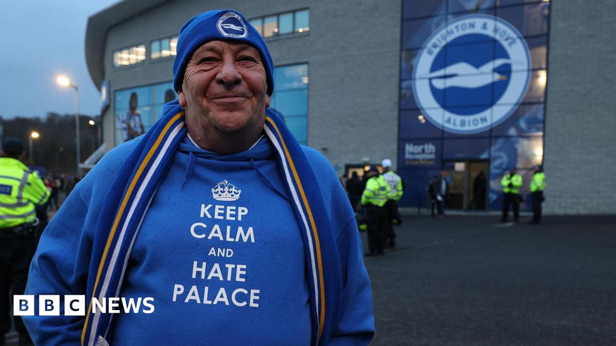 A man in a Brighton and Hove Albion beanie and hoodie that says "Keep calm and hate Palace". He is standing in front of a Brighton badge on the outside of the Amex.