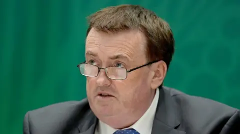 PA Media A man with short brown hair, metal glasses and blue eyes, wearing a black suit, white shirt and blue tie.