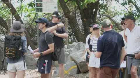 Men and women wearing shorts and T-shirts stand on sand with rocks, a tree and a sign behind them.