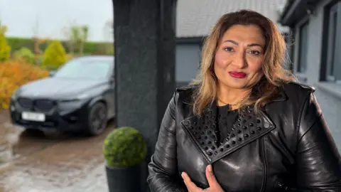 A woman in leather jacket sits in a wheelchair looking into the camera on her front porch. In the background a black car is parked in the driveway.
