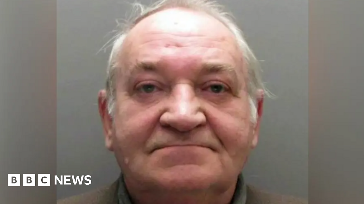 Peter Toole standing next to a black door. He has short grey hair and blue eyes. He is wearing a cream long-sleeved top with a small black writing on it.