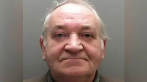 Durham Police A mugshot of Neville Husband. He has short white hair and dark eyes.  He is wearing a dark blue turtleneck top and a grey suit jacket. 