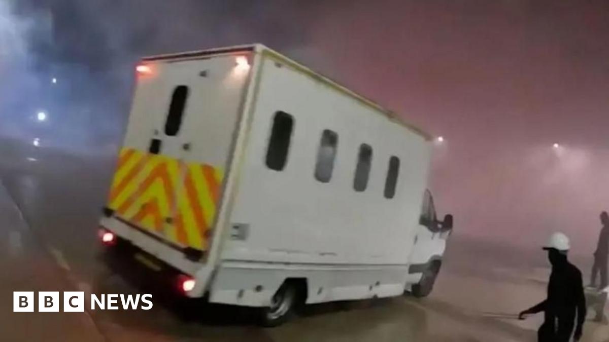 A white van with yellow and orange hi-vis decals at night. There is a lot of smoke in the air and on the right there is a figure wearing all black and a white hard hat.