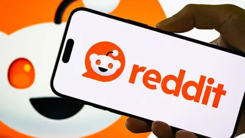 Getty Images A hand holding a mobile phone with the reddit logo with the reddit character in the background