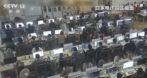 CCTV Screenshot from a CCTV documentary showing security camera footage of a scam centre, with workers sitting at rows of chairs, each looking at their computer screens.