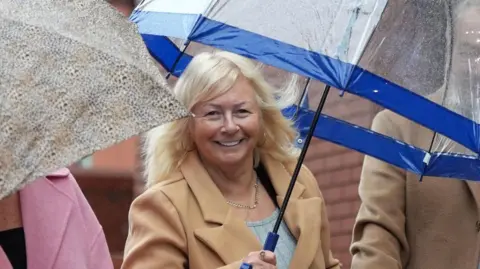 PA Media Mrs Spragg wearing a tan jacket and holding an umbrella standing next to two people either side of her