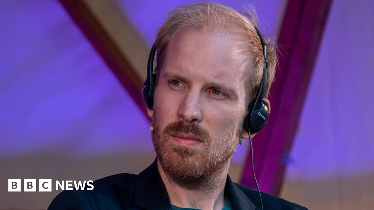 Rutger Bregman, wearing headphones, is looking to his right with a thoughtful expression - he is wearing a dark suit that can just be seen, he has blond-red hair and a short beard and moustache - he is inside a building with purple lighting behind him