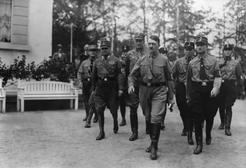 General Photographic Agency/Hulton Archive/Getty Images Black and white image of Hitler walking along with uniformed men behind him