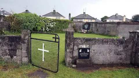 Getty Images There is a stone wall courtyard with grass growing in it, where the former mother-and-baby home stood. A black metal gate with a white cross attached to a stone wall has swung open in the picture. There are bushes and flowers growing in the courtyard, and there is a plaque at the gate remembering those that died there.