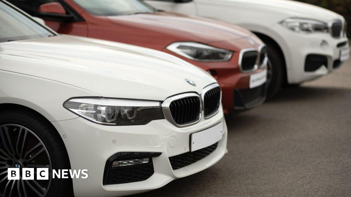 A close-up shot shows three BMW cars parked in a diagonal row on a paved surface. The front car is white with a prominent grille and headlights, while a red BMW sits behind it, followed by another white BMW.