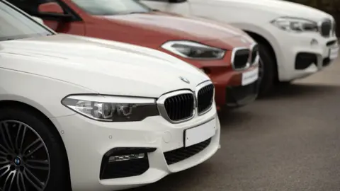 Getty Images A close-up shot shows three BMW cars parked in a diagonal row on a paved surface. The front car is white with a prominent grille and headlights, while a red BMW sits behind it, followed by another white BMW.