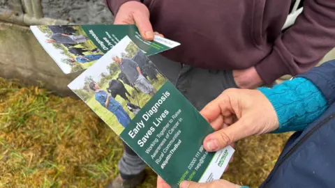 Two flyers are being held by two sets of hands. The flyer is headlined 'early diagnosis saves lives'. In the background you can see that the people holding. the flyers are dressed warmly in boots and standing on grassy ground. 