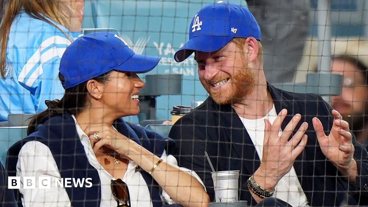 Meghan Markle and Prince Harry wear blue Dodgers baseball caps while seated in the front row during the third inning between the Toronto Blue Jays and the Los Angeles Dodgers during Game 4 of the 2025 MLB World Series at Dodger Stadium.