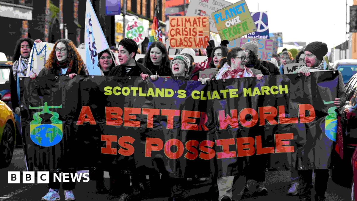 A large group of protesters marches down a street holding a wide black banner that reads “Scotland’s Climate March” and “A Better World Is Possible” in bold coloured letters. People at the front appear to be chanting, while others behind them hold signs with messages such as “Climate Crisis = Class War,” “Planet Over Profit,” and a peace symbol. The crowd is tightly packed, and buildings line the street in the background.