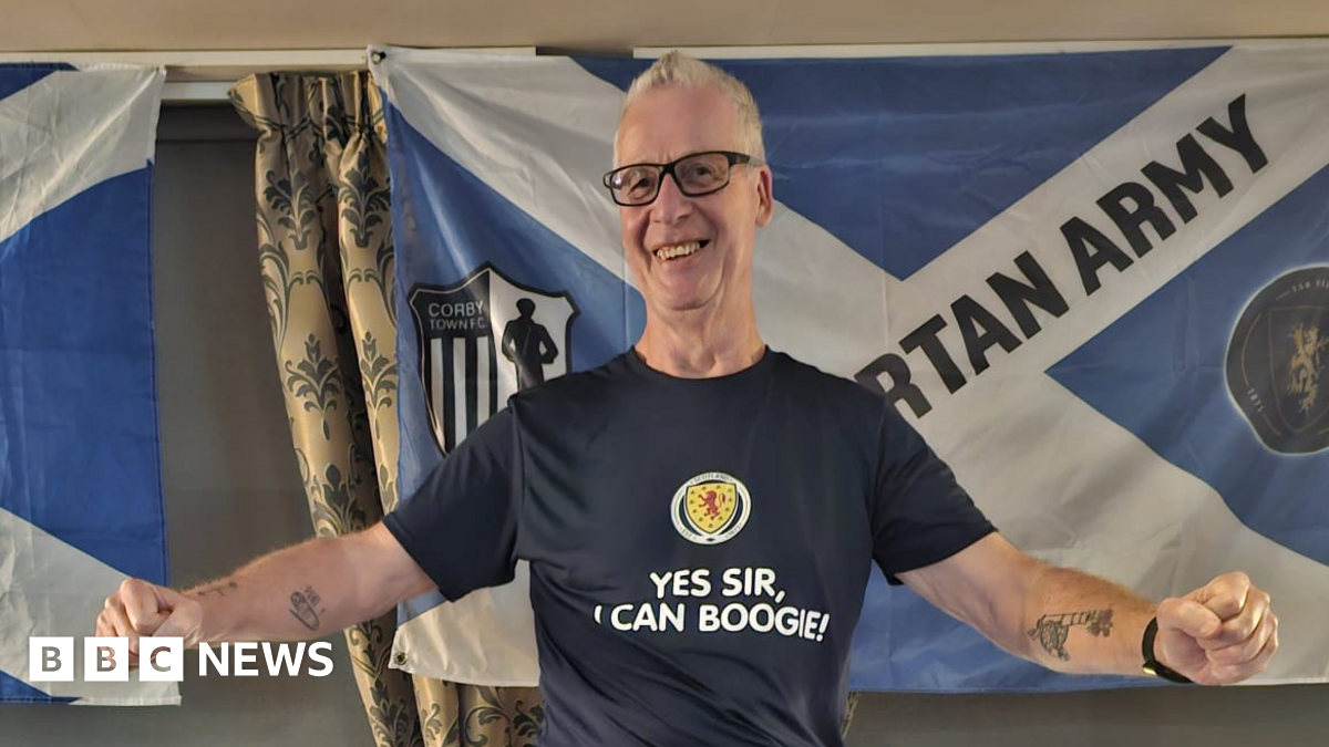 A man in glasses and a navy T-shirt stands in front of two Scotland flags, smiling. He is wearing glasses and a dark blue shirt which has the Scotland badge on it and the words "Yes sir, I can boogie!"