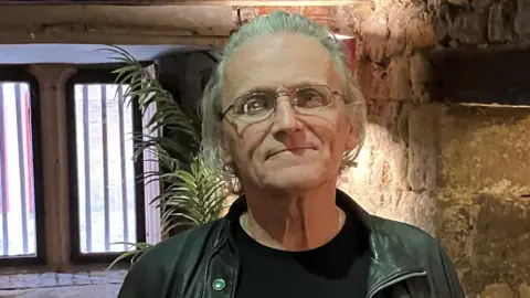 Jimmy Coffey is standing in front of a window, next to a stone wall. His grey hair is swept back. He is wearing glasses, a black top and a black leather jacket. 