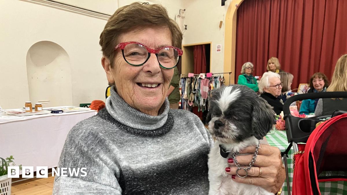 A woman in red framed glasses holding a small grey dog on her lap. She is sitting in a village hall