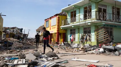 EPA/Shutterstock Two men look among the rubble of a street. Shops can be seen behind them partially damaged