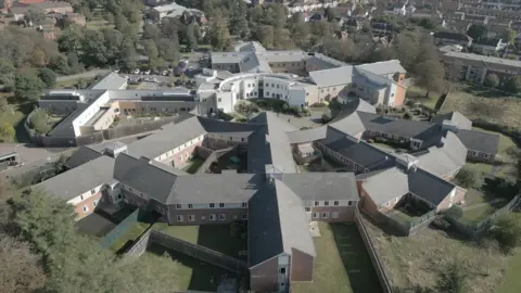 An aerial photograph shows Goodmayes Hospital from above. Buildings span out from a central circular area. It is surrounded by grass and trees.