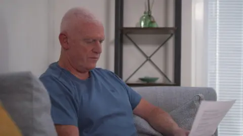 BBC Paul Harrison has short grey hair and is wearing a blue t-shirt. He is sitting on a grey sofa in his living room and reading a letter.