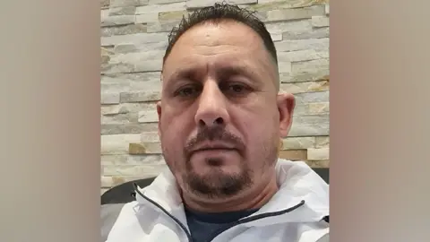 TikTok Robert Calin, a man with short dark hair and a goatee beard, wearing a grey and white tracksuit top and pictured in close-up against a brick wall