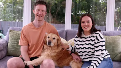 British Heart Foundation A man and woman sit on a grey couch, with a golden retriever dog between them. The man wears an orange t-shirt and black short. The woman wears blue jeans and a black and white striped jumper. Behind them are clear windows with white frames. Outside are green trees and a black fence, separating the trees from their garden 